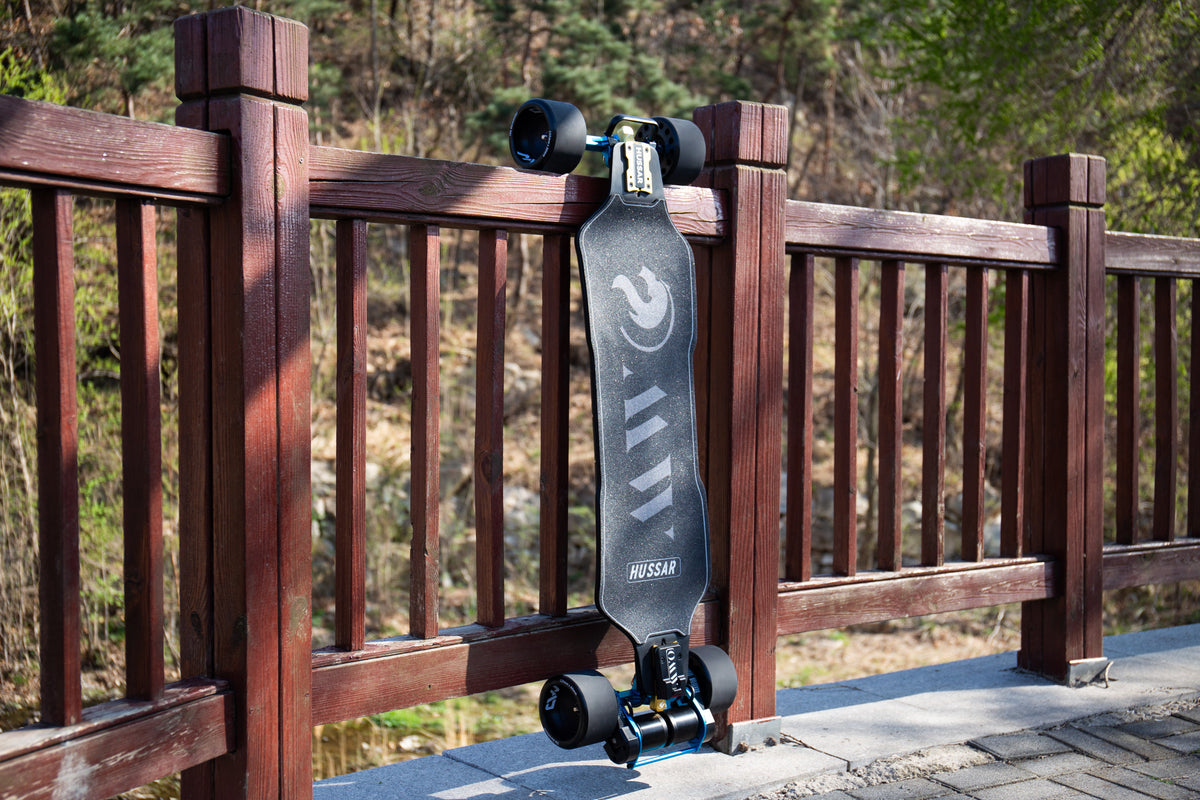 OMW Hussar electric skateboard with 125 mm MAD MAX wheels and TKP trucks beside a wooden railing on a dry mountain route